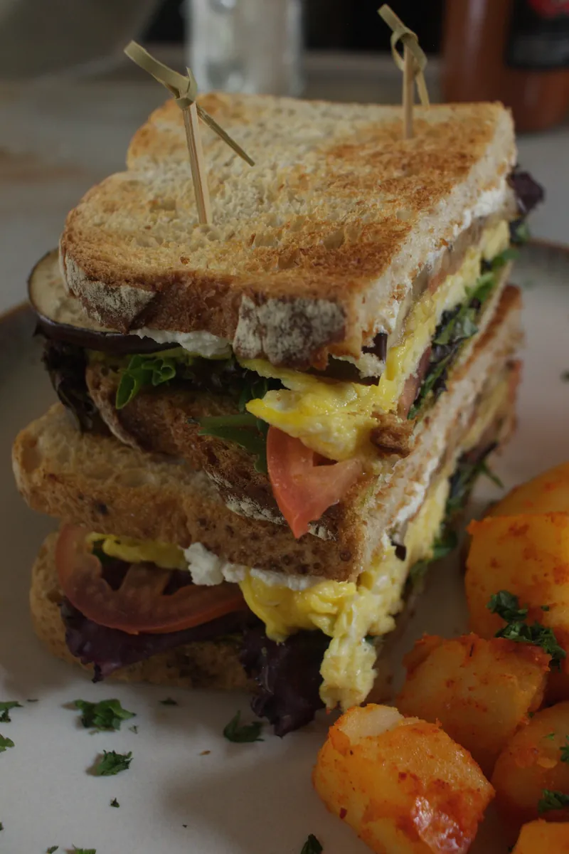 Triple-decker breakfast club sandwich with egg, tomato, greens, and goat cheese on toasted sourdough with home fries