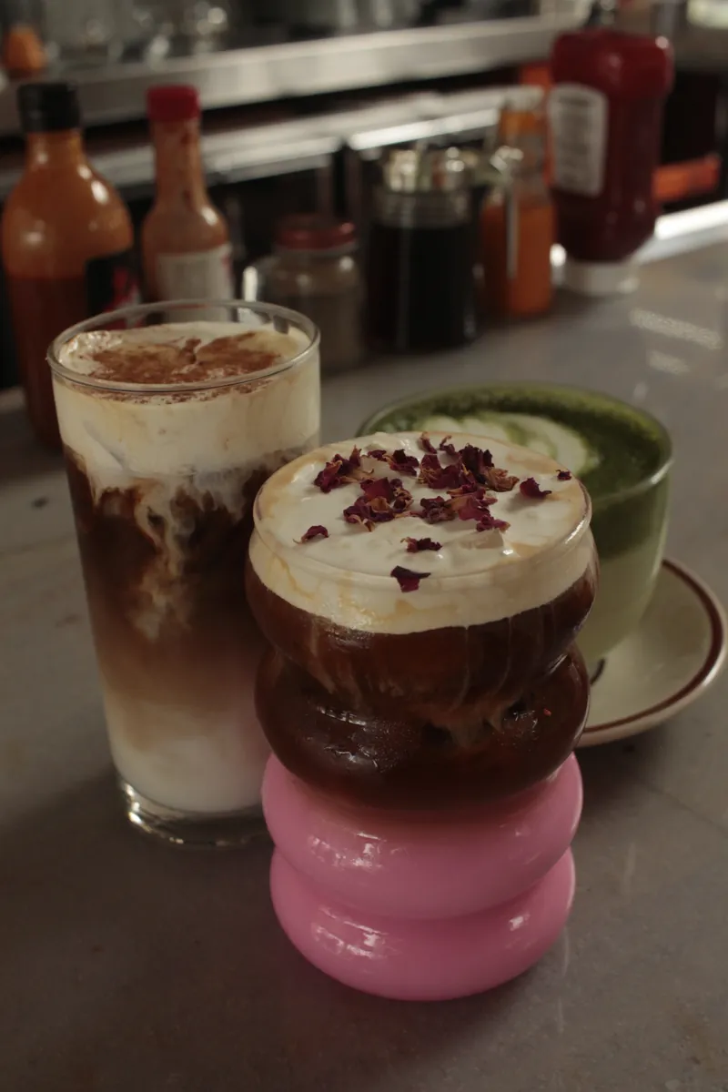 A flight of specialty drinks on the bar — rose petal iced coffee, cinnamon-dusted latte, and a matcha